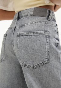 Jean en denim gris à taille haute, présentant un tissu texturé, deux poches arrière et une écusson en cuir de marque à la ceinture.