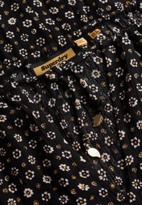 Zwarte jurk van stof met bloemenprint, versierd met kleine witte bloemen en gele accenten, met een geplooide halslijn en decoratieve knopen.