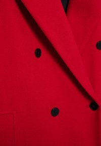 Červený vlnený dvojradový kabát s čiernymi gombíkmi, s výrezovým golierom a jemnou textúrou. Obsahuje detail bočného vrecka.