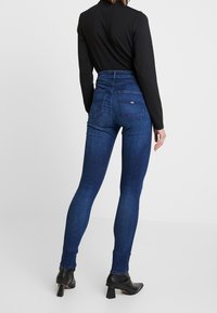 Mujer vestida con jeans skinny oscuros de talle alto con bolsillos traseros, camiseta negra de manga larga y botas de tacón negras hasta el tobillo, de pie sobre un suelo blanco.