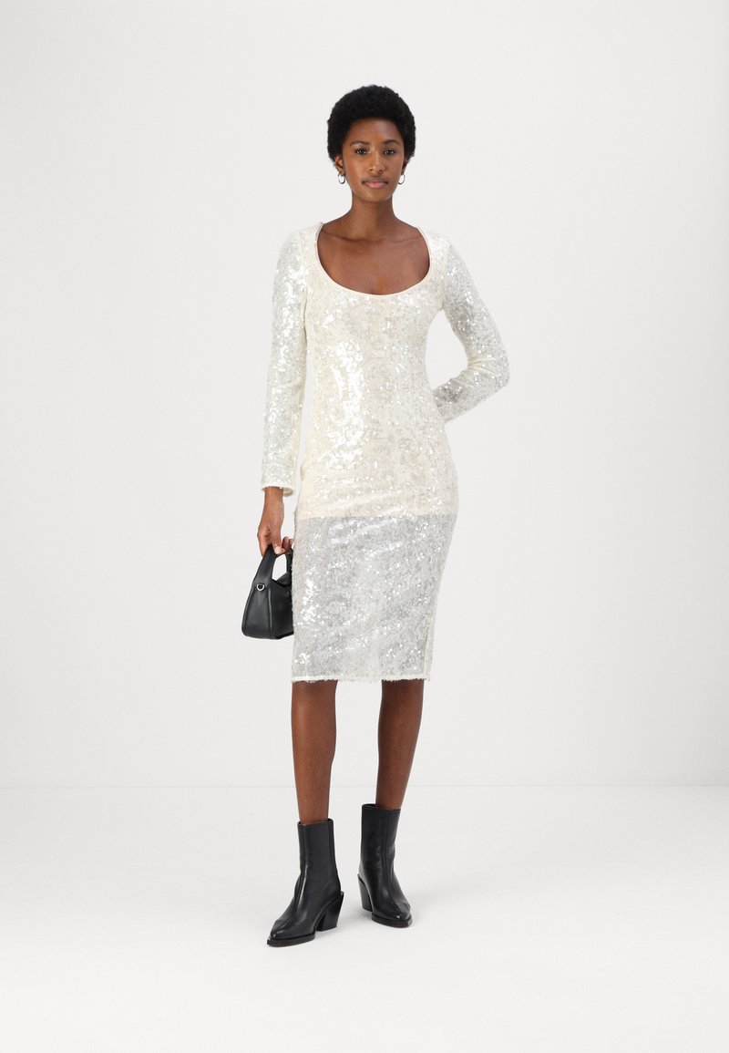 Sukienka z cekinami, z długimi rękawami, dekoltem w serek, dopasowanym fasonie, przechodząca od kremowego do srebrnego, zestawiona z czarnymi botkami za kostkę i małą torebką.