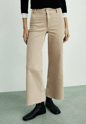 Osoba nosi beżowe spodnie z wysokim stanem i szerokimi nogawkami z postrzępionymi brzegami oraz czarne botki, stoi na tle jasnego, jednolitego tła.