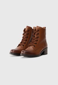 Botas de tobillo de cuero marrón con cordones en la parte delantera, puntera redondeada, suela de goma y un tacón modesto. Presentan una lengüeta en la parte posterior para ponerlas y quitarlas fácilmente.