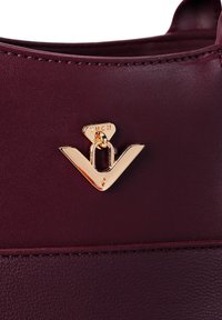 Burgunderfarbene Kunstledertasche mit einem goldfarbenen Metallverschluss in Form eines Dreiecks und einer glatten Textur. Sichtbare Steppnähte.