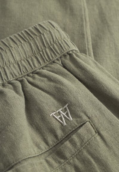 Primer plano de pantalones de tela verde con cintura elástica y las iniciales "AA" bordadas en blanco sobre la costura de un bolsillo.
