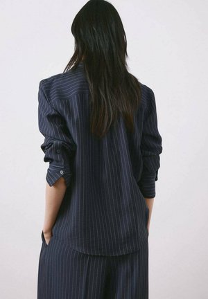 Persona con cabello largo y oscuro, vistiendo una camisa azul marino con rayas finas y pantalones anchos a juego, con las manos en los bolsillos, de espaldas.