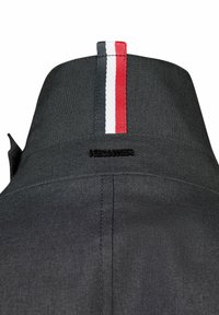 Zwarte jas met een kraag en een gestreepte lus in rood, wit en blauw stof. Heeft een logo in zwart geborduurd op de achterkant. Gestructureerd materiaal.