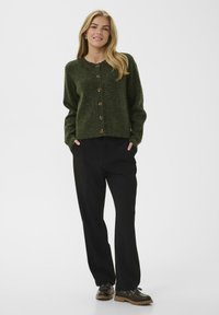 Tumši zaļš vilnas džemperis ar apaļu kakla izgriezumu, septiņiem pogu aizdari, ribotām piedurknēm un brīvu piegulumu, kombinēts ar melnām platām biksēm.