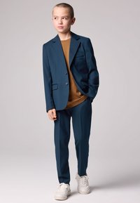 Sinivihreä puku, jossa yksirivinen bleiseri avoimilla taskuilla ja siihen sopivat kapealahkeiset housut, päällä ruskea paita ja yhdistetty valkoisiin lenkkareihin.