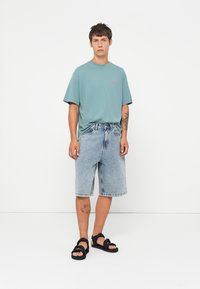 Young man standing wearing a teal oversized T-shirt, light blue denim knee-length shorts, black sandals, with tattoos on arm and leg.
