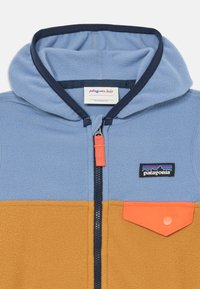 Flisasta zip-up kapuca z modrim zgornjim delom, oranžnim spodnjim delom in usklajeno žepom. Ima mornarsko obrobe in logo Patagonia.