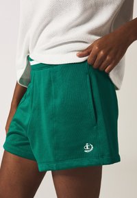 Pantalones cortos verdes de un material suave, con bolsillos laterales, una cinturilla blanca en contraste y un pequeño logo bordado en el dobladillo.