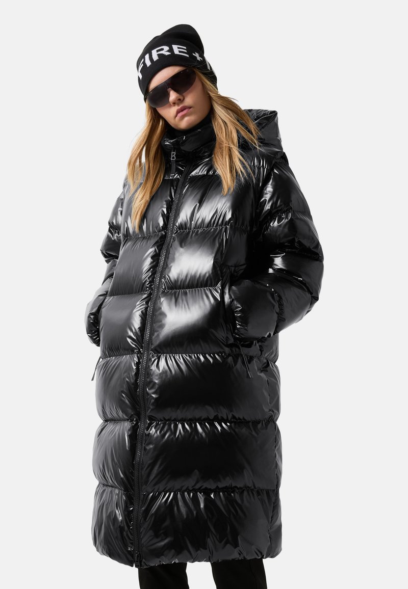 Vrouw in een lange glanzende zwarte pufferjas met capuchon, zwarte beanie met witte tekst, donkere zonnebril, handen in de zakken, op een lichtgrijze achtergrond.