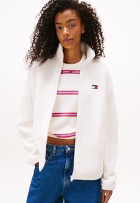 Witte fleece zip-up jacket met een hoge kraag, gecombineerd met een gestreepte cropped sweatshirt in rood en blauw, en blauwe spijkerbroek.