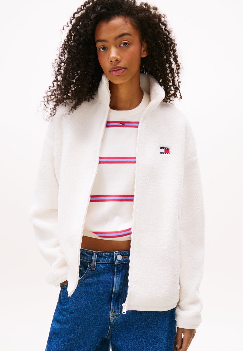 Witte fleece zip-up jacket met een hoge kraag, gecombineerd met een gestreepte cropped sweatshirt in rood en blauw, en blauwe spijkerbroek.