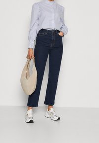 Chemise à rayures avec manches bouffantes, jeans bleu foncé, baskets blanches avec accents gris et sac à main en suède beige.