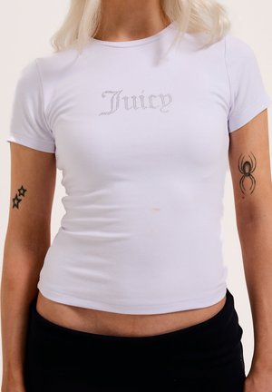 Weißes eng anliegendes T-Shirt mit Strassschriftzug "Juicy", getragen von einer Person mit Stern- und Spinnentattoos an den Armen vor einfarbigem Hintergrund.