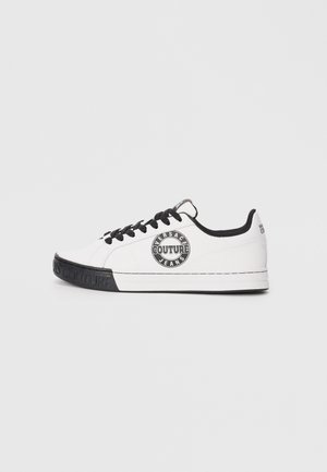 Hvit sneaker med svarte detaljer, med en rund logo-patch på siden, svarte snørebånd, strukturert materiale og gummisåle.