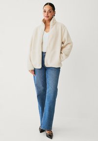 Cremefarvet fleecejakke med høj krave, lynlåsclosure og forlommer, parret med blå lige ben jeans og sorte spidse sko.