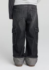 Pantaloni cargo in denim nero con vestibilità ampia, dotati di due tasche laterali e orlo ripiegato in un tessuto grigio a contrasto. Tasche posteriori visibili.
