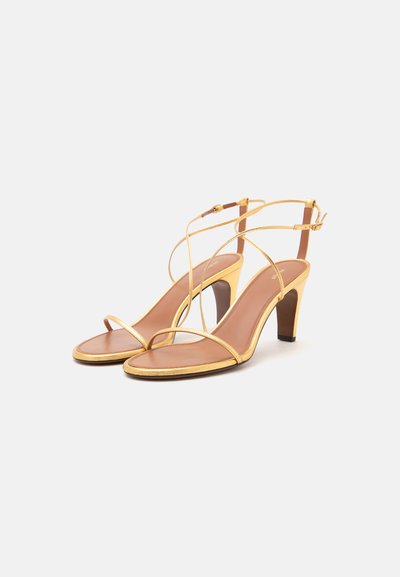Paire de sandales dorées à talons hauts avec fines brides à la cheville et orteils ouverts sur un fond clair uni.