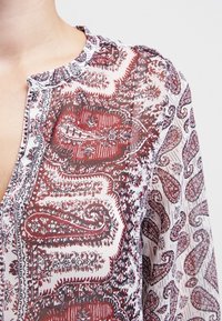 Durchsichtige Bluse mit roten und floralen Paisleymustern, strukturiertem Stoff, Stehkragen und V-Ausschnitt. Besitzt aufwendige Details im gesamten Design.