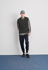Grønn ribbestrikket vest over en stripet skjorte, kombinert med marineblå joggebukser og svarte sko. En lys blå caps fullfører antrekket mot en nøytral bakgrunn.