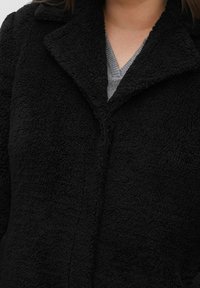 Musta, teksturoitu fleecetakki lovekulmaisella kauluksella ja edessä napillinen suljenta, joka tarjoaa mukavan ja lämpimän designin.