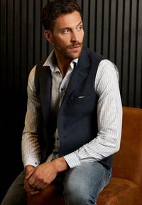 Männliches Modell trägt eine dunkle Weste über einem hell gestreiften Hemd, sitzt mit verschränkten Händen. Hintergrund zeigt dunkle vertikale Linien.