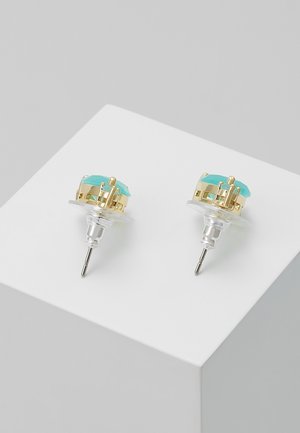 Een paar oorknopjes met gouden zettingen en lichtblauwe gefacetteerde stenen, tentoongesteld op een witte ondergrond.