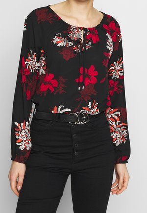 Černá halenka s dlouhým rukávem s červenými a bílými květinovými vzory, uvázaným výstřihem, doplněná o černé kalhoty s vysokým pasem a černý pásek s dvojitými kroužky.