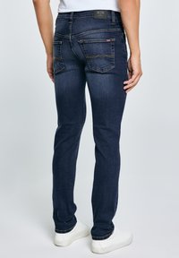 Osoba ubrana w dopasowane ciemnoniebieskie dżinsy i białe trampki, pokazana od tyłu na jednolitym jasnym tle.