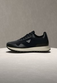 Baskets basses noires avec logo blanc sur le côté, lacets noirs et semelle beige avec une semelle texturée, présentées sur une surface et un fond neutres.
