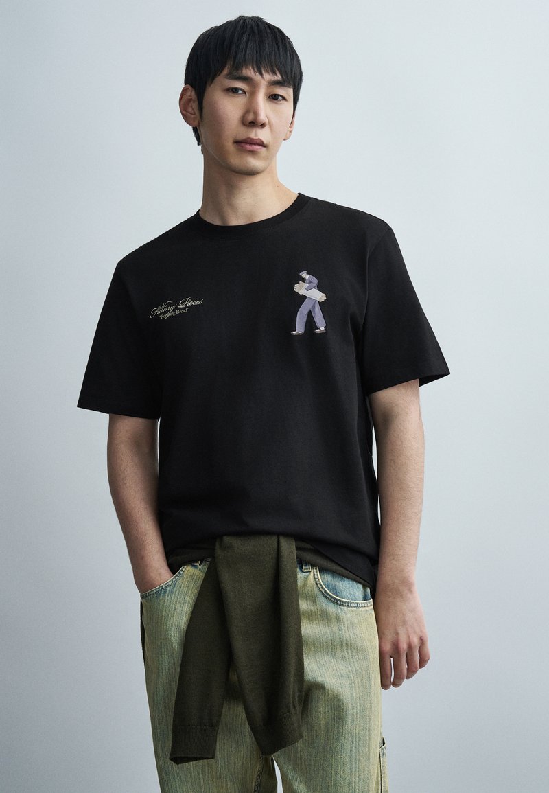 Μαύρο βαμβακερό μπλουζάκι με μικρό κεντητό σχέδιο μιας φιγούρας και κείμενο στο αριστερό στήθος, σε συνδυασμό με ανοιχτό μπλε τζιν παντελόνι και πράσινο πουλόβερ.