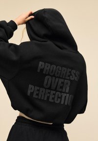 Črna skrajšana jopa s kapuco, na zadnji strani z mat besedilom "NAPREDEK PRED POPOLNOSTJO," zasnovana za udobje.