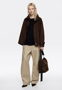 Chaqueta ligera marrón, suéter negro y pantalones amplios beiges. El modelo sostiene un bolso marrón con una correa. Materiales texturizados, corte relajado.