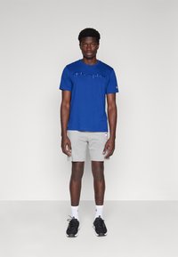 Camisa de tirantes azul con el logotipo de "Champion" en cursiva, combinada con pantalones cortos gris claro y zapatillas deportivas negras. Calcetines blancos visibles.