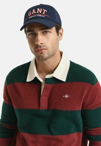 Gestreifter Pullover in Grün und Burgunderrot mit einem weißen Kragen und gesticktem Logo, dazu eine blaue Kappe mit roten Schriftzügen.