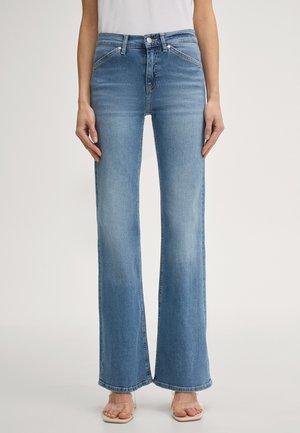 Frau trägt hellblaue, hoch taillierte Schlagjeans und offene Absatzschuhe mit transparentem Riemen, steht vor einem schlichten Hintergrund.