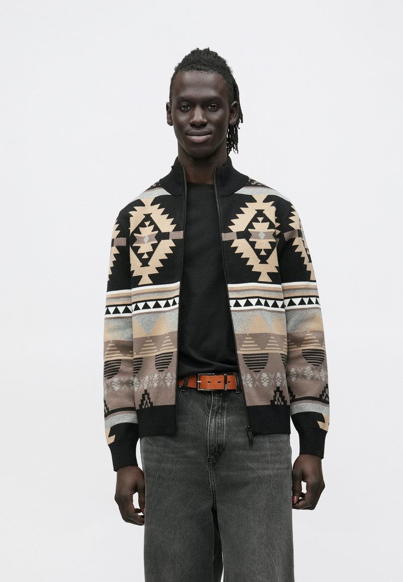 Mladi moški s temno poltjo, ki nosi črn pulover in kardigan z zadrgo, okrašen z geometrijskimi vzorci v barvah tan, bež in črni, stoji pred enobarvnim ozadjem.