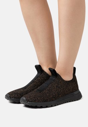 Baskets noires sans lacets en tissu pailleté avec languettes, portées aux pieds sur un fond clair uni.