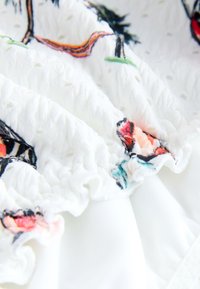 Tela blanca con volantes texturizados y bordado floral colorido, que presenta tonos de rosa, rojo y verde sobre un fondo suave.