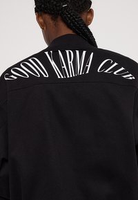 Czarna kurtka z napisem "GOOD KARMA CLUB" w pogrubionych, białych literach na plecach. Materiał wygląda na gładki, o dopasowanym kroju.