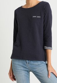 Navy blue polka dot long-sleeve top with a round neckline, featuring a small chest pocket and striped accents on the sleeve cuffs.