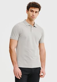 Jasnoszara koszulka polo z klasycznym kołnierzykiem, krótkimi rękawami, zapięciem na dwa guziki i dopasowanym fasonem. Noszona z czarnymi spodniami.