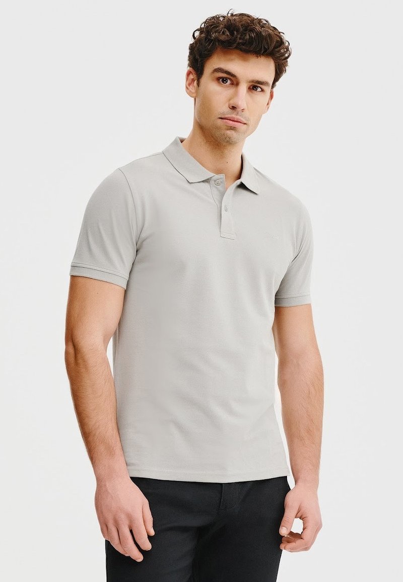 Jasnoszara koszulka polo z klasycznym kołnierzykiem, krótkimi rękawami, zapięciem na dwa guziki i dopasowanym fasonem. Noszona z czarnymi spodniami.