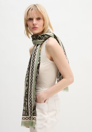 Donna bionda che indossa un top bianco senza maniche e pantaloni bianchi, con una sciarpa verde e nera a motivo avvolta intorno al collo.