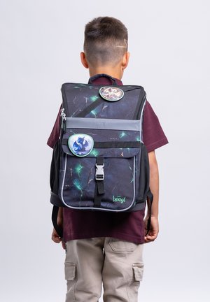 Boy wearing dark patterned backpack decorated with two round cartoon dragon badges, standing against plain background, facing away.