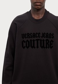 Crna dukserica s okruglim izrezom i raglan rukavima. Sadrži istaknuti crni logo s tekstom "VERSACE JEANS COUTURE" u podebljanom fontu.