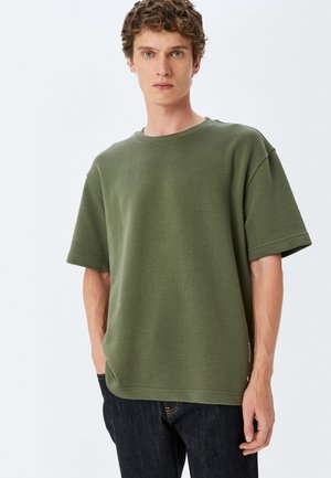 Oliven grøn kortærmet t-shirt i en afslappet pasform, med rund hals og subtile sømdetaljer, parret med mørke jeans.